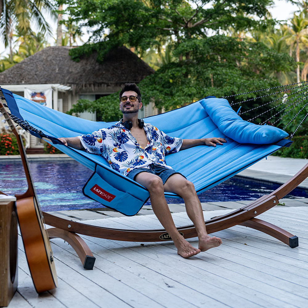 Man relaxing on Lazy Daze Double Hammock with Curved Spreader Bar in blue by the backyard pool.#color_blue