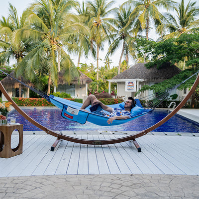 A man resting on Lazy Daze Double Hammock with Curved Spreader Bar in blue by the pool.#color_blue