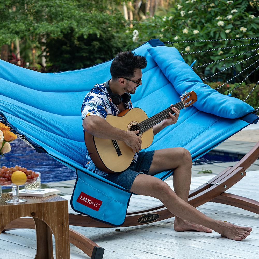 Man relaxing in Lazy Daze Double Hammock with Curved Spreader Bar in blue, enjoying the backyard.#color_blue