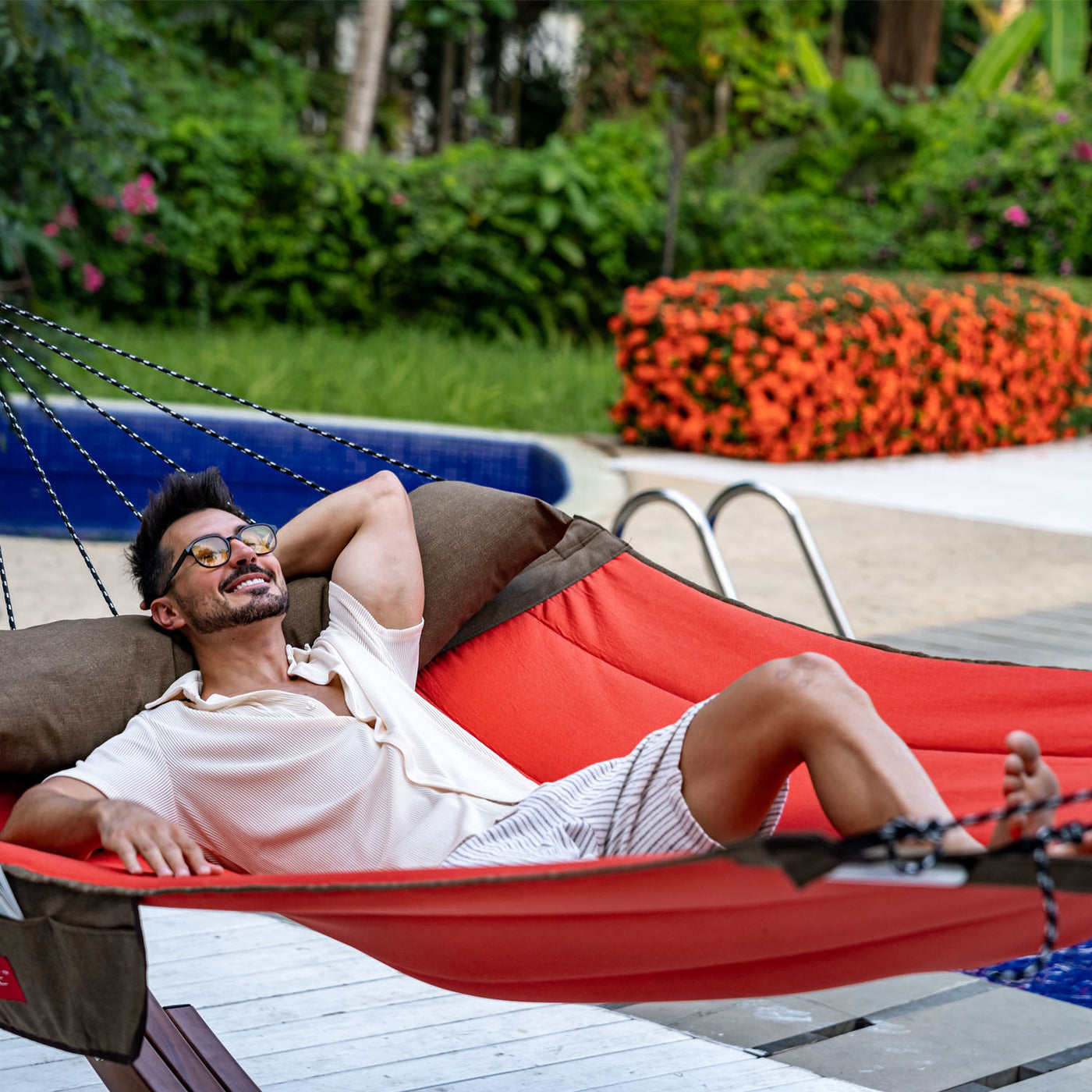 A man relaxing in the Lazy Daze Double Hammock with Curved Spreader Bar in red, set in a peaceful backyard.#color_red