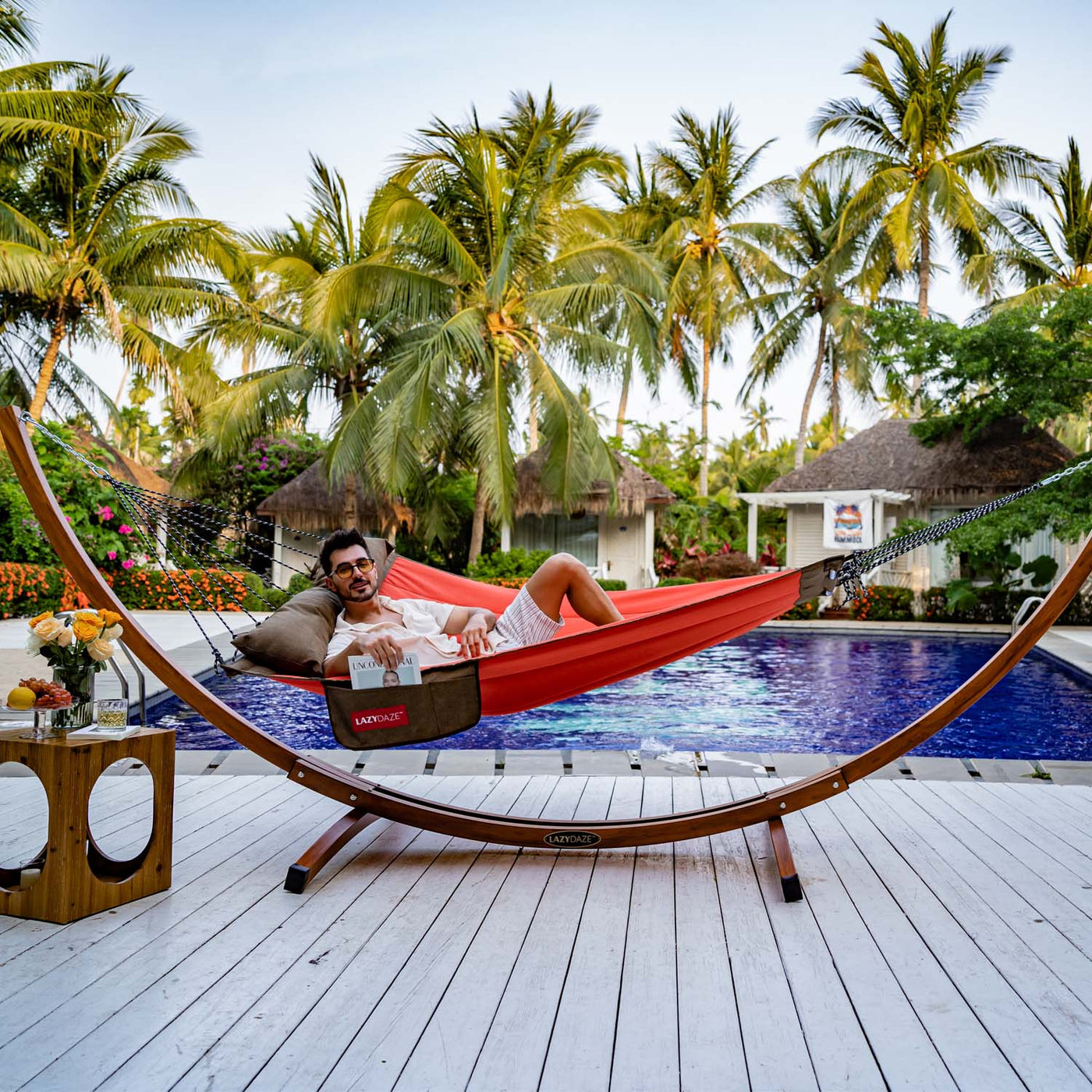 A man lounging in the Lazy Daze Double Hammock with Curved Spreader Bar in red, surrounded by lush greenery.#color_red