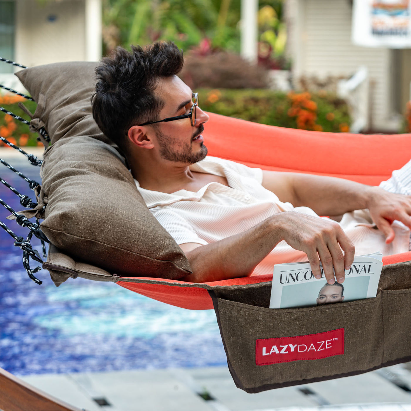 A man reclining in the Lazy Daze Double Hammock with Curved Spreader Bar in red, enjoying a sunny backyard.#color_red