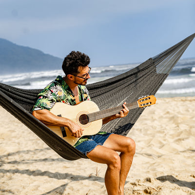 Close-up of a man resting in the Lazy Daze Mayan Family Hammock in gray on the sandy shore.#color_gray