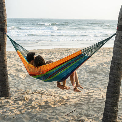 A couple relaxing on the Lazy Daze Mayan Family Hammock in rainbow color at the beach.#color_multi-color