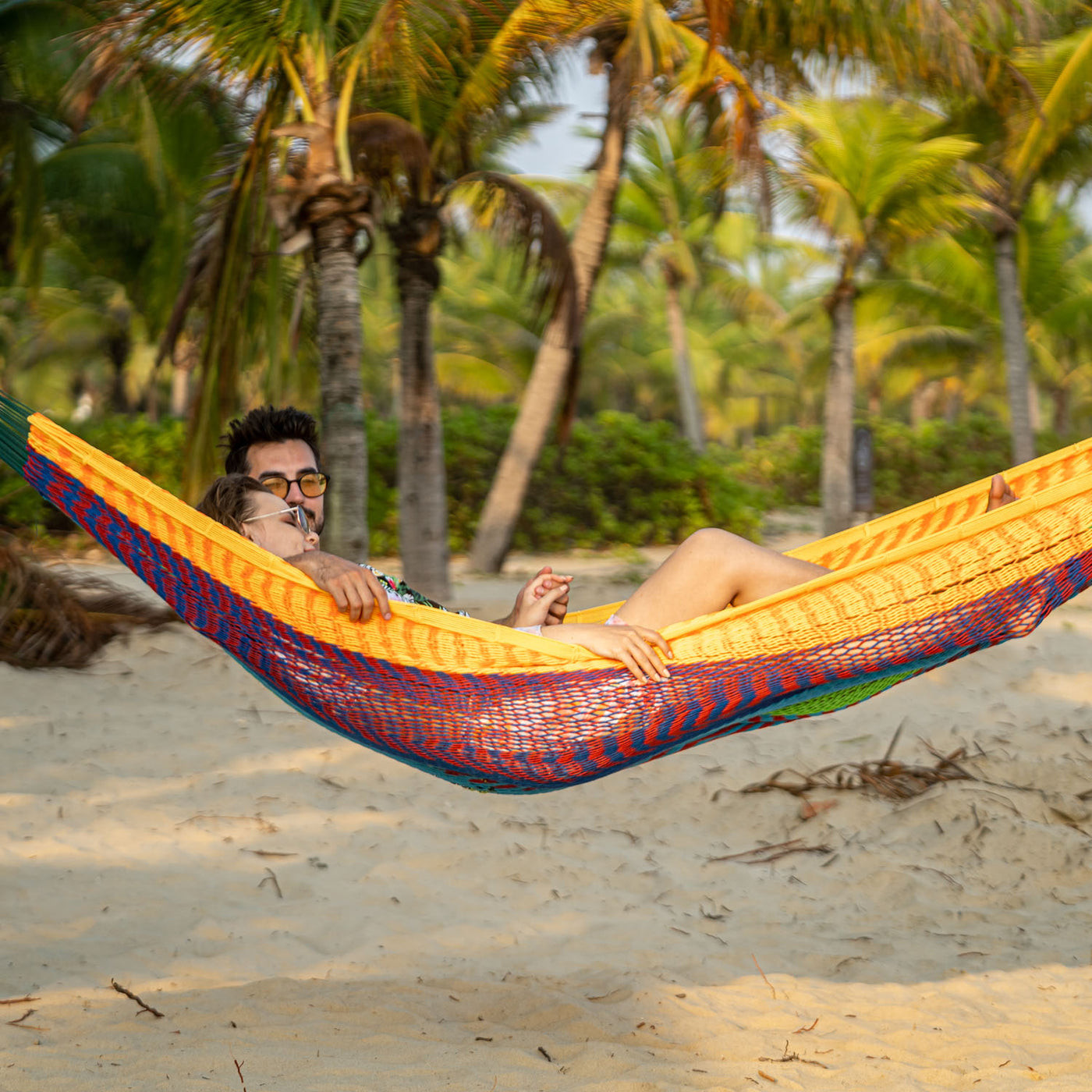 A man and woman enjoying the Lazy Daze Mayan Family Hammock in rainbow under a shady tree on the beach.#color_rainbow