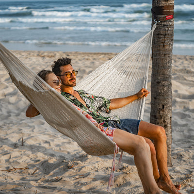 A man and woman reclining on the Lazy Daze Mayan Family Hammock in white near the ocean.#color_white